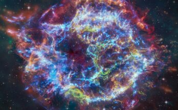 a giant shell of wispy blue, green, yellow and red light glows against the blackness of space in a photo by space telescopes.
