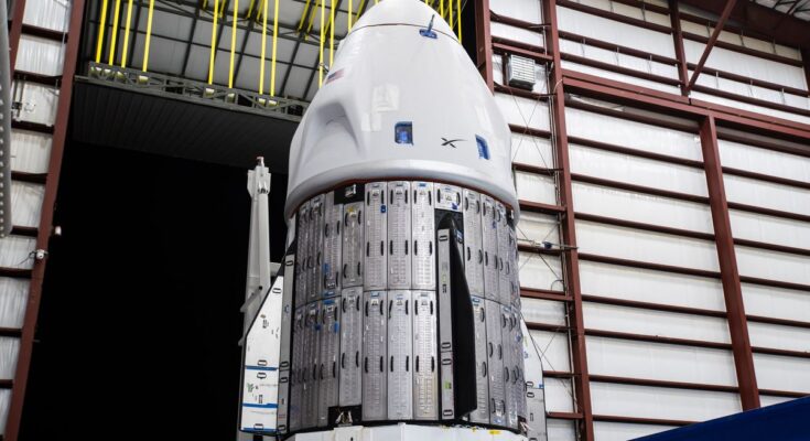 a white spacex dragon capsule is seen inside a large hangar near a door that opens onto darkness.