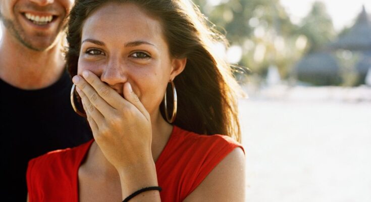 Teenage girl with brunette hair, wearing a red top and gold hoop-earrings is shown giggling with her left hand over her mouth. Behind her is a man who is wearing a black t-shirt. The top half of his face is cut off. The background is blurry but the faint outline of trees can be seen.