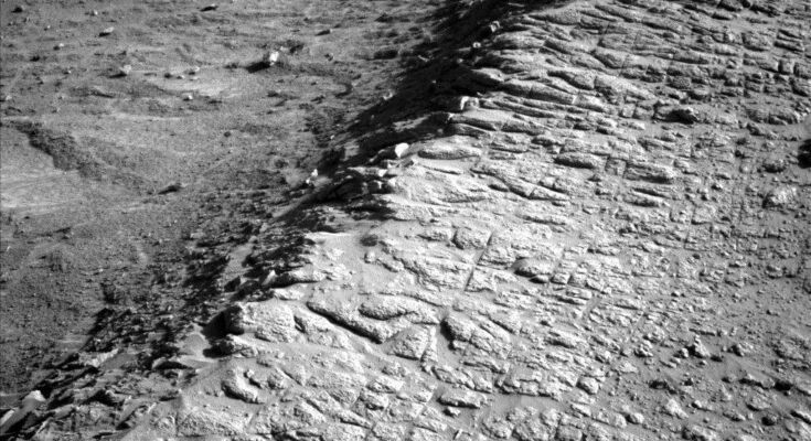 A grayscale photo from the Martian surface shows rough terrain covered in small rocks, divided across the middle of the image by a ridge running from the lower left corner of the frame to the upper right corner. The ground to the right of the ridge is relatively flat, while to the left it falls away from the ridge into a depression. The entire scene is light to medium gray, except for a very dark shadow along the left side of the ridge.