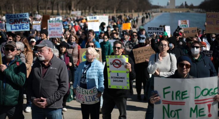 People holding signs rally in front of the Washington Monument in DC. One sign reads "Science Saves Lives"; another says "Fire DOGE, not me"