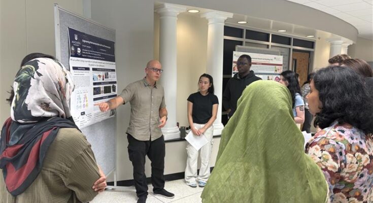 A man in a grey shirt and glasses, Dr. Paul Langridge, presents a research poster to a crowd of PhD students.