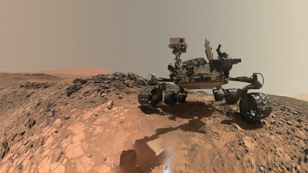 Un rover es un montañoso debajo del cielo gris plano, sentado en la superficie del naranja Marte, con un trozo de roca a su alrededor.
