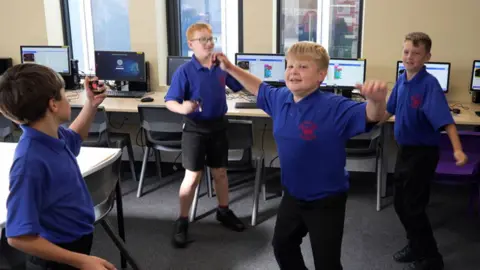 John Fair Hall/BBC cuatro años 6 niños usan polo azul brillante y pantalones y pantalones cortos negros. Están bailando en el aula. Detrás de ellos hay una larga línea de mesas, cada una en un monitor de computadora y un teclado y sillas frente a ellas. 