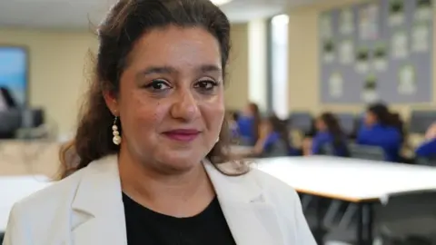 John Fair Hall/BBC Shahnila Saeed, se retiró de la frente con cabello largo negro. Ella comienza a sonreír con una chaqueta de crema en la parte superior negra. Ella está de pie en el aula, está borrosa, pero puedes encontrar mesas y estudiantes en GA Bright Blue Stations. 
