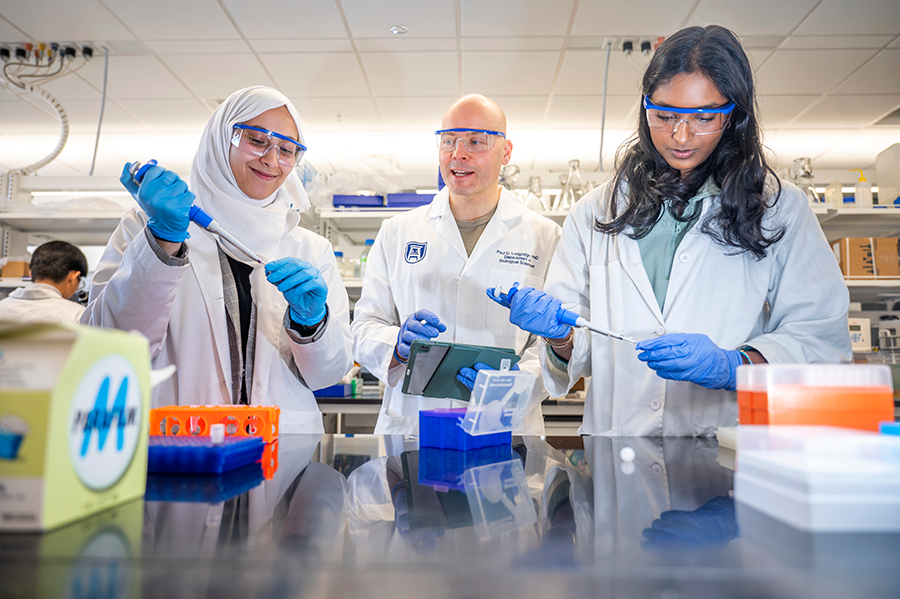 El científico masculino está parado en el laboratorio entre dos científicas. Todos usan guantes, gafas de seguridad y abrigos de laboratorio.