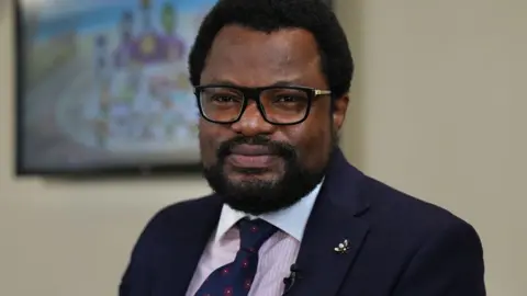 John Fair Hall/BBC Jude Enjemke con cabello negro corto y una pequeña barba negra. Lleva una corbata azul, así como una chaqueta azul marino sobre gafas de marco negro, camisa rosa y blanca. Él se para en la pared con una débil pantalla detrás de él. 