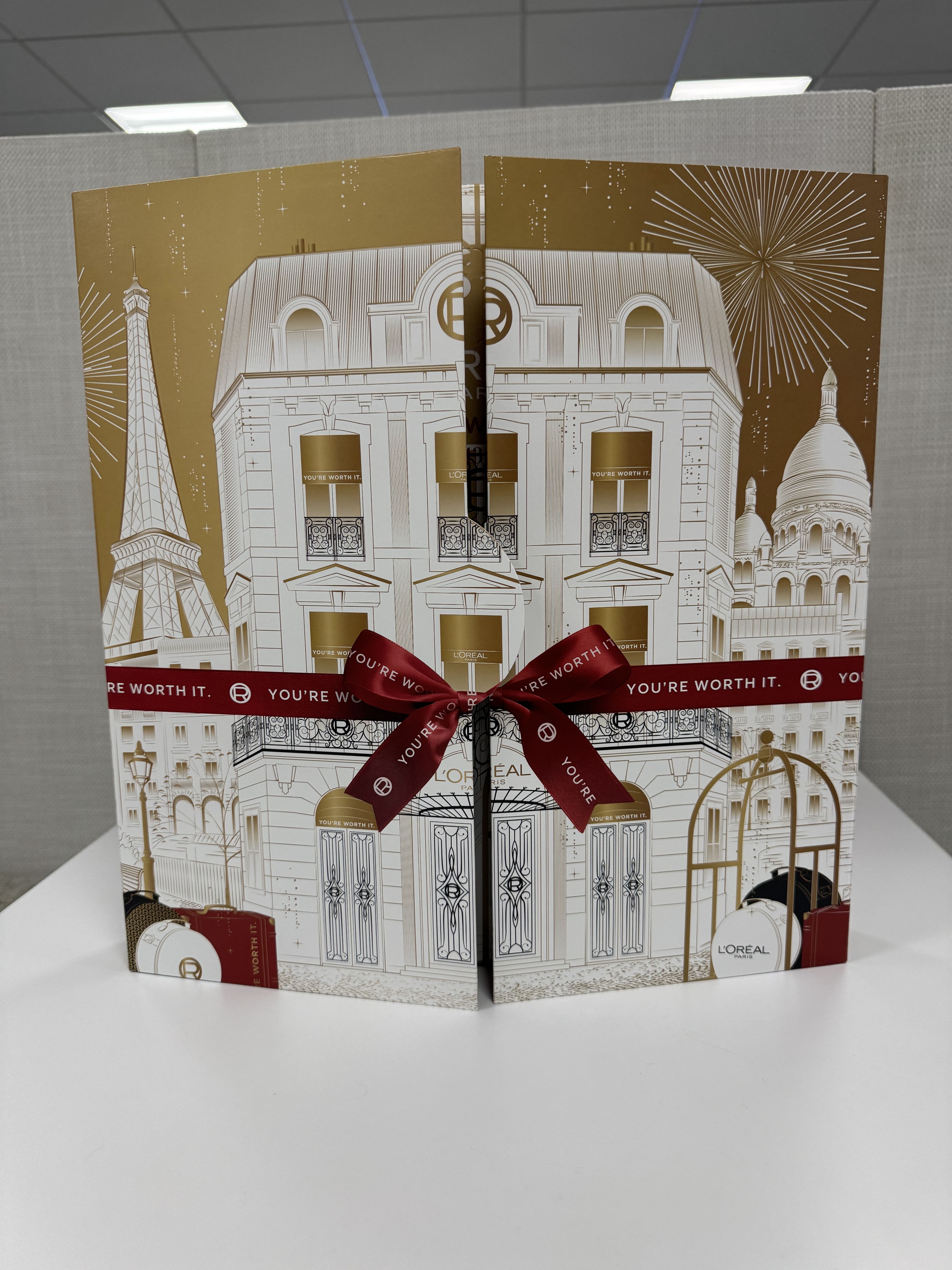 Una caja de regalo de L'Oréal con una representación de una escena callejera de París que incluye la Torre Eiffel y el Sacré-Coeur, atada con una cinta color burdeos.