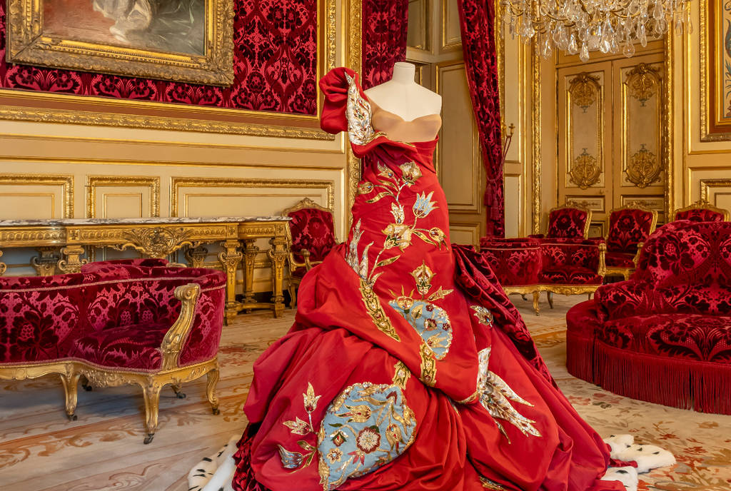 La imagen muestra un vestido de terciopelo rojo dentro de una sala decorada para el Louvre Couture en el Museo del Louvre.