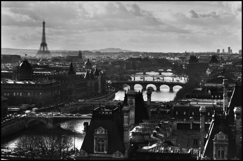 Una vista en blanco y negro de París bajo un cielo nublado con la Torre Eiffel a lo lejos, varios puentes que cruzan el río Sena y edificios históricos de la ciudad en primer plano.