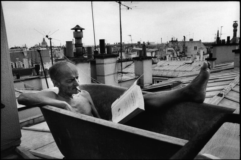 Un anciano se relaja en una vieja bañera en el techo, leyendo un libro con una pierna en alto. El horizonte de la ciudad, las chimeneas y las antenas se ven al fondo bajo un cielo nublado.
