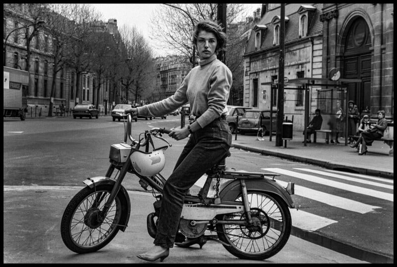 Una mujer con un suéter y jeans sentada en un ciclomotor en una intersección de calles de la ciudad y mira a la cámara. Al fondo se ven árboles sin hojas y varias personas sentadas en un banco.
