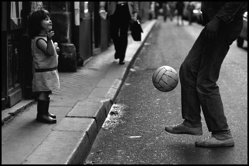 Un niño pequeño está parado en una acera, sonriendo y mirando a un adulto patear un balón de fútbol por la calle en una escena urbana en blanco y negro. Los peatones caminan al fondo.