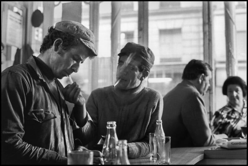 Dos hombres se sientan en el mostrador de una cafetería; Uno enciende el cigarrillo del otro. Ambos visten ropa de trabajo y gorras. En el mostrador hay botellas de vidrio y vasos vacíos. Al fondo, otros dos padres sentados junto a la ventana conversan.