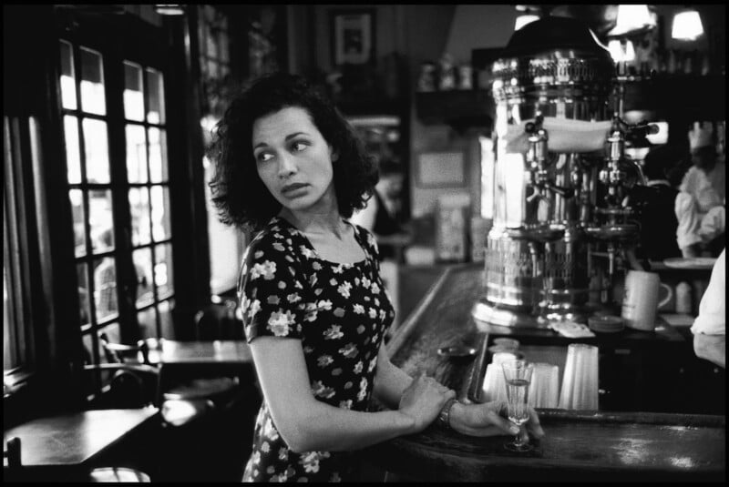 Una mujer de pelo oscuro y rizado con un vestido de flores se encuentra en un bar, mirando hacia la izquierda con expresión pensativa. Hay un vaso vacío sobre el mostrador y el fondo muestra el interior de una cafetería de aspecto antiguo.