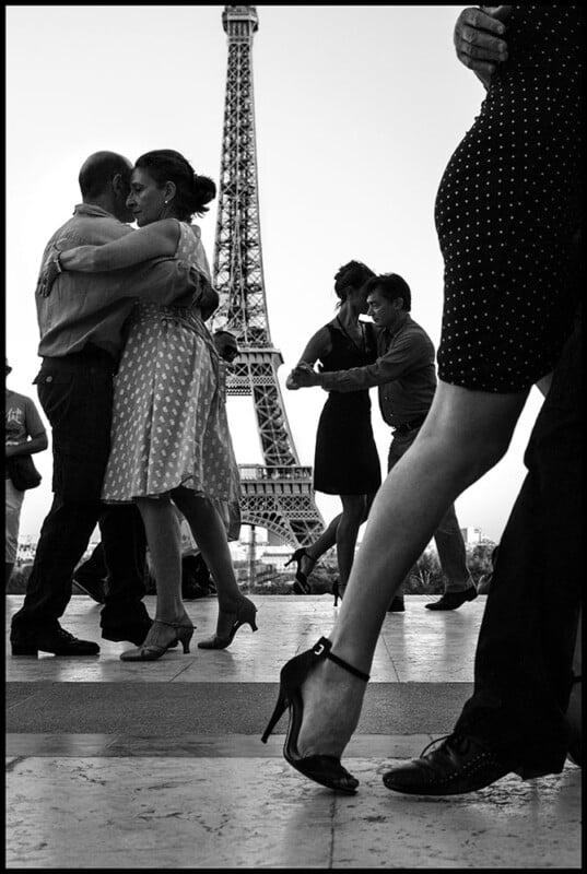 La pareja baila íntimamente frente a la Torre Eiffel. La foto es en blanco y negro y muestra a personas con ropa elegante y tacones altos, resaltando el movimiento y el romance en un ambiente parisino.