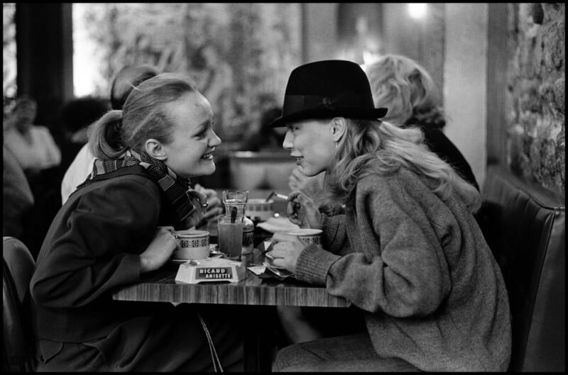 Dos mujeres están sentadas juntas en una mesa de café, sonriendo e inclinándose la una hacia la otra para conversar. Ambos tienen bebidas y cuencos, uno lleva una bufanda y el otro un gorro y un suéter holgado. Otros padres están al fondo.