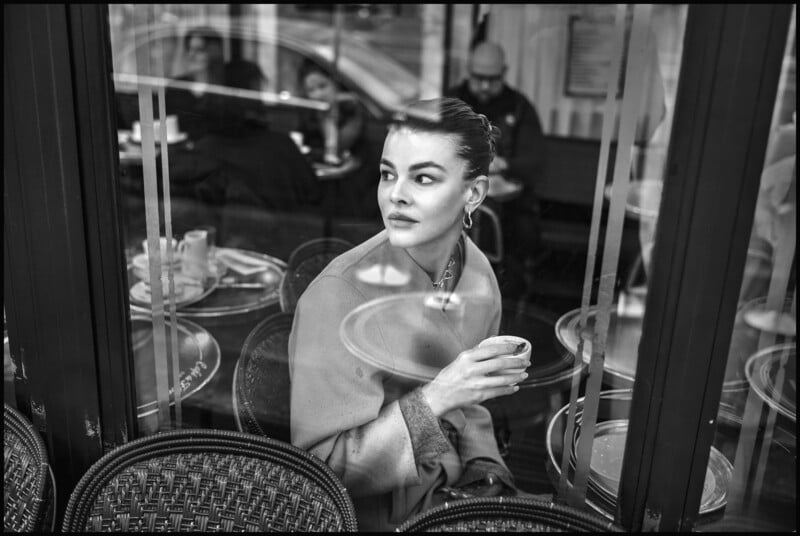 Una mujer sentada en una mesa de café junto a la ventana, sosteniendo una taza pequeña y mirando pensativamente hacia un lado. Hay varias tazas y platos vacíos sobre las mesas a su alrededor, con otras personas visibles al fondo. La foto está en blanco y negro.