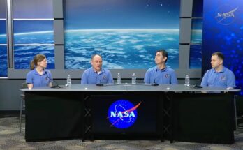 Four individuals wearing pale blue shirts sit behind a long table with a black cloth over it and the blue NASA circle logo with blue screens behind them.