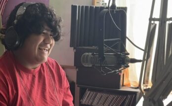 Person recording a podcast in a studio, wearing headphones and smiling, with a microphone and sound equipment.